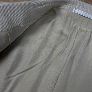 May include: Close-up of the interior lining of a light beige blazer. The lining is a smooth, dotted fabric, with visible seams and a textured collar. The blazer's interior is visible, showing the construction and materials.