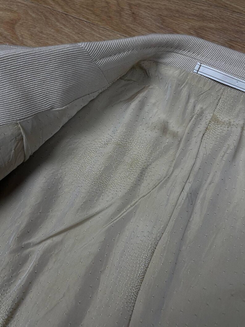 May include: Close-up of the interior lining of a light beige blazer. The lining is a smooth, dotted fabric, with visible seams and a textured collar. The blazer's interior is visible, showing the construction and materials.