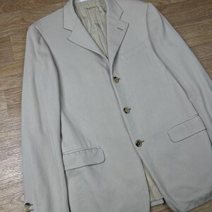 May include: A light beige blazer with a notched lapel, three-button closure, and two front pockets. The blazer has four buttons on each cuff and a light beige lining. The garment is made of a textured fabric.