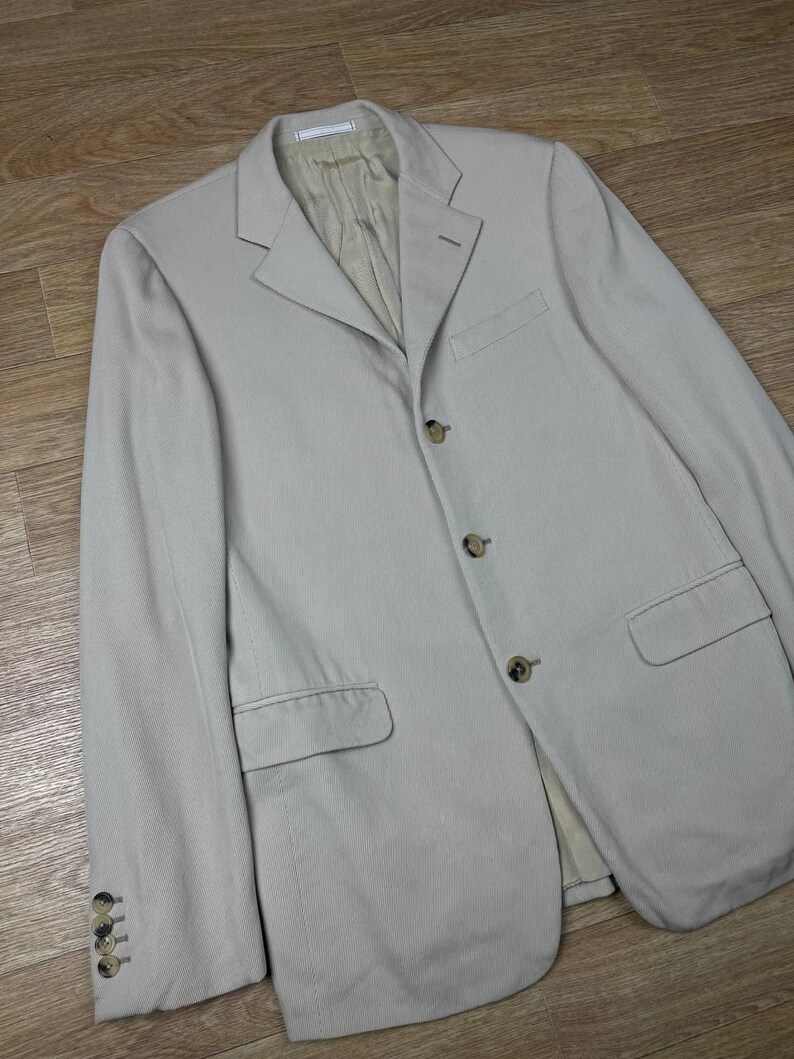 May include: A light beige blazer with a notched lapel, three-button closure, and two front pockets. The blazer has four buttons on each cuff and a light beige lining. The garment is made of a textured fabric.