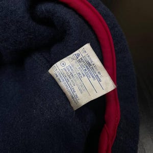 May include: A dark navy blue fleece garment with a bright red trim along the edge. A white care instruction label is sewn onto the interior, featuring text in English and Italian that includes washing, bleaching, ironing, and dry cleaning symbols.
