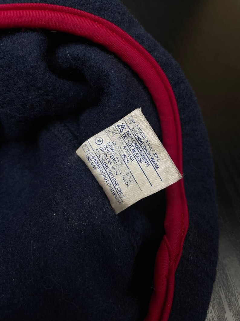 May include: A dark navy blue fleece garment with a bright red trim along the edge. A white care instruction label is sewn onto the interior, featuring text in English and Italian that includes washing, bleaching, ironing, and dry cleaning symbols.