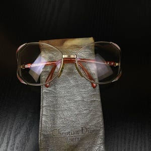 May include: Gold-tone metal eyeglasses with large, rounded frames and reddish-brown temple arms, resting on a silver-colored textured fabric pouch. The pouch features the embossed text Christian Dior on the lower front section of the fabric.