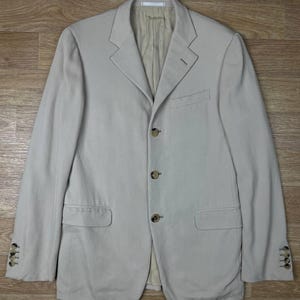 May include: A light beige blazer with a three-button closure and two front pockets. The blazer has a notched lapel and buttoned cuffs. The jacket is made of a light-colored fabric and is laid flat on a wooden surface.