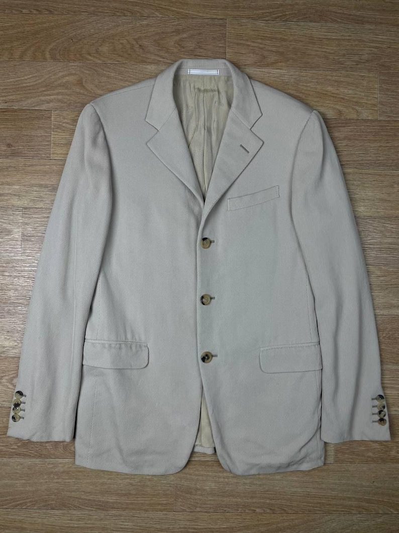 May include: A light beige blazer with a three-button closure and two front pockets. The blazer has a notched lapel and buttoned cuffs. The jacket is made of a light-colored fabric and is laid flat on a wooden surface.