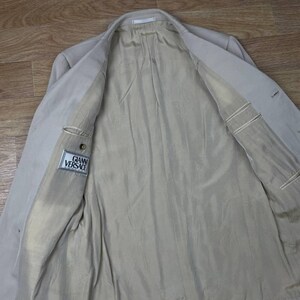 May include: Beige blazer with a light-colored lining. The inside of the jacket is visible, showing pockets and the "GIANNI VERSACE" label. The blazer is open, revealing the interior construction and fabric.