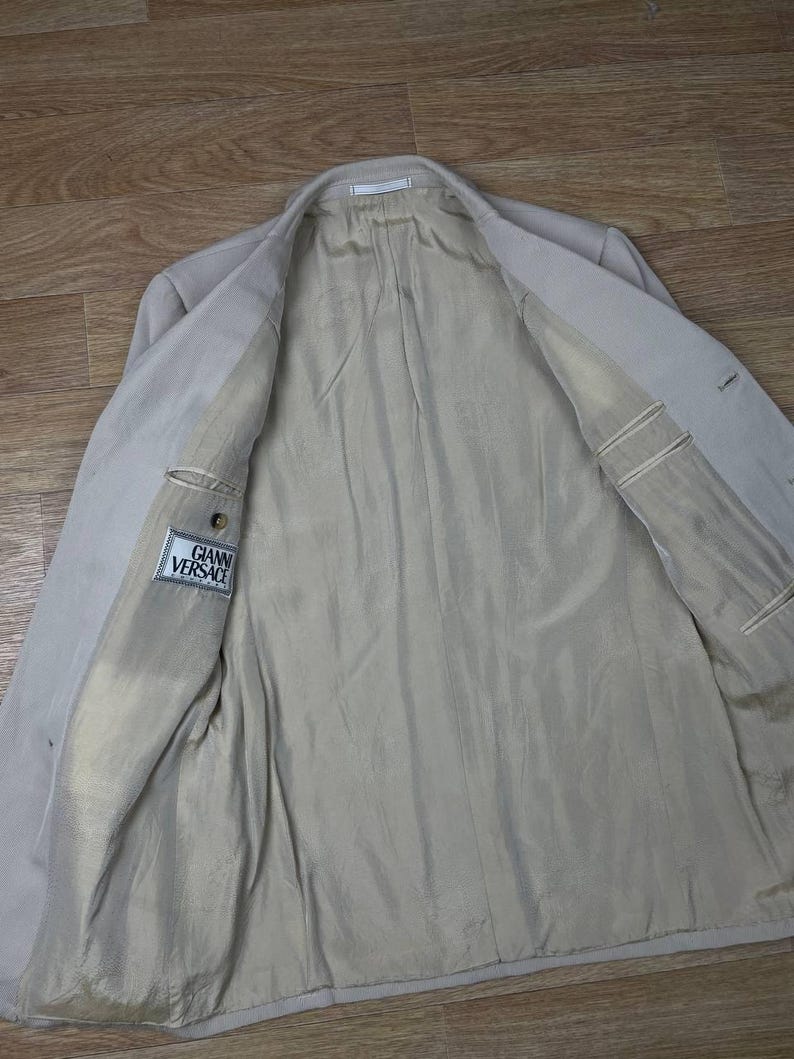 May include: Beige blazer with a light-colored lining. The inside of the jacket is visible, showing pockets and the "GIANNI VERSACE" label. The blazer is open, revealing the interior construction and fabric.
