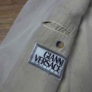 May include: Close-up of a beige blazer interior with a pocket and a button. The Gianni Versace Couture label is visible. The fabric has a textured, dotted pattern and a ribbed section.