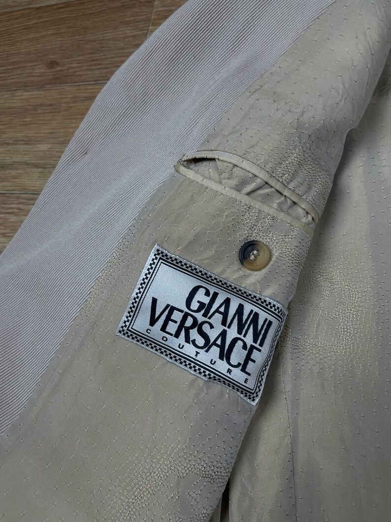 May include: Close-up of a beige blazer interior with a pocket and a button. The Gianni Versace Couture label is visible. The fabric has a textured, dotted pattern and a ribbed section.