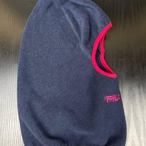 May include: A navy blue fleece balaclava featuring a bright pink trim around the face opening and a pink embroidered logo on the side. The headgear is constructed from a soft, textured fabric and includes a contoured hood design.