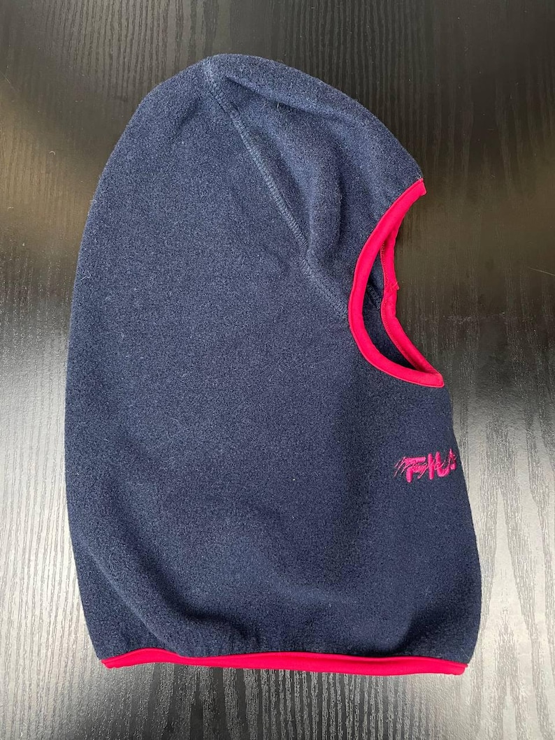 May include: A navy blue fleece balaclava featuring a bright pink trim around the face opening and a pink embroidered logo on the side. The headgear is constructed from a soft, textured fabric and includes a contoured hood design.