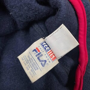 May include: A navy blue fleece garment with a bright red trim along the edge. A white fabric tag is attached, featuring the Fila logo in blue and red, the text 'tee tech', 'ONLY FROM', '100% POLYESTER', and 'ISTRUZIONI SUL RETRO FOR CARE SEE OVER'.