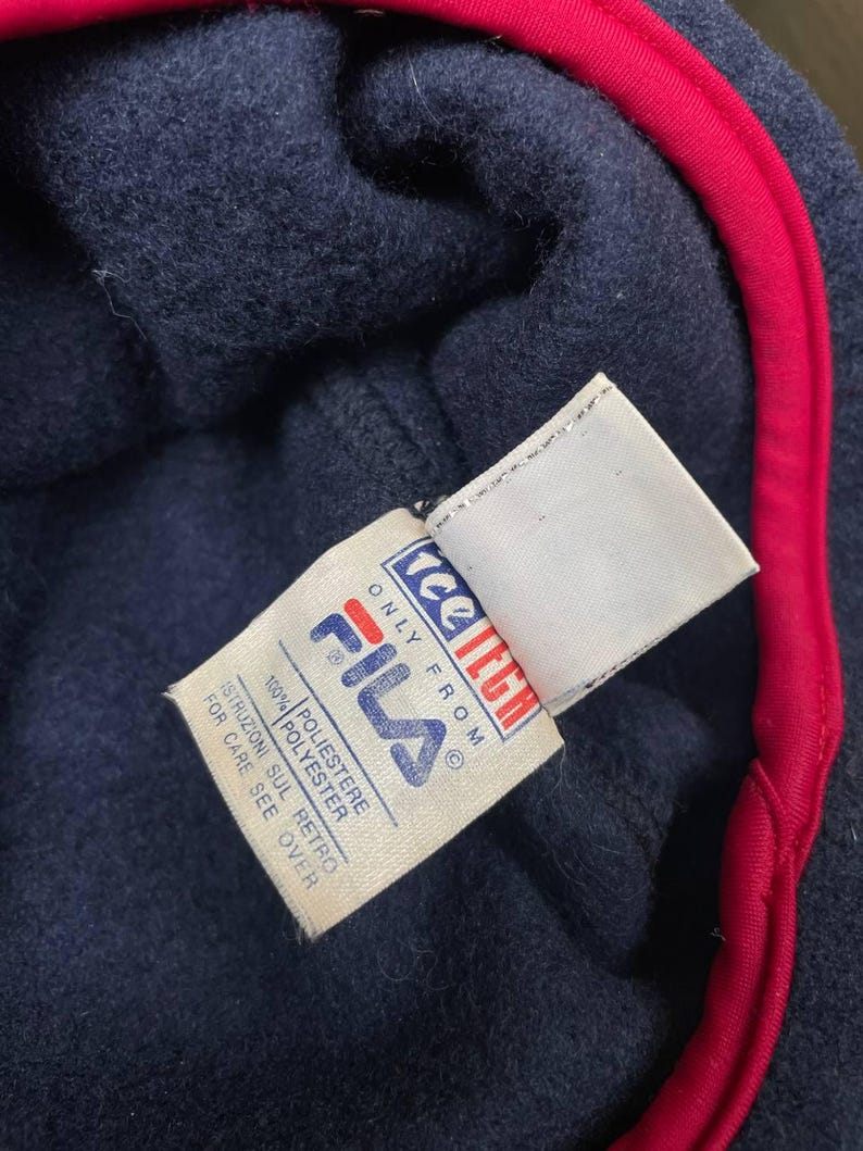 May include: A navy blue fleece garment with a bright red trim along the edge. A white fabric tag is attached, featuring the Fila logo in blue and red, the text 'tee tech', 'ONLY FROM', '100% POLYESTER', and 'ISTRUZIONI SUL RETRO FOR CARE SEE OVER'.