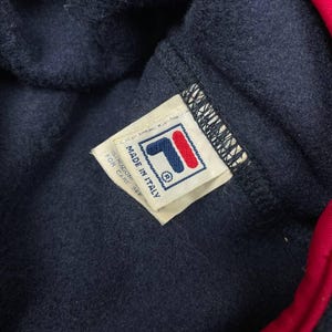 May include: A navy blue fleece garment with a bright red trim along the edge. A white fabric tag is stitched to the interior, featuring the Fila logo in blue and red, with the text Made in Italy and Istruzioni per care see printed on it.