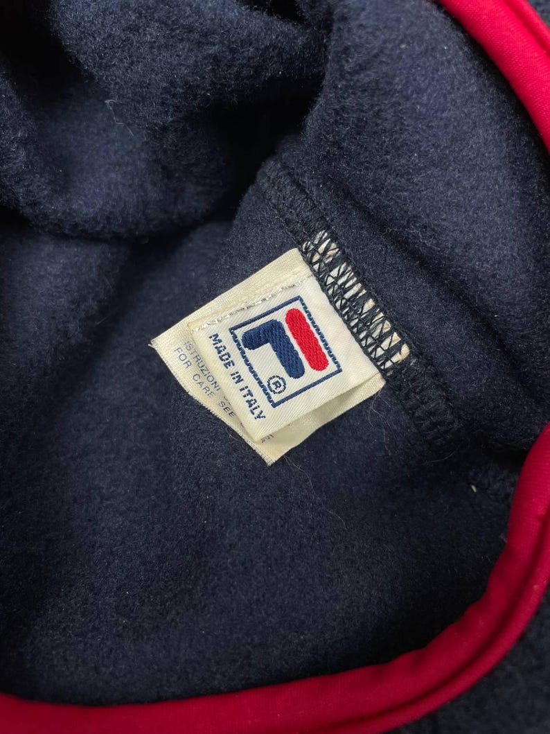 May include: A navy blue fleece garment with a bright red trim along the edge. A white fabric tag is stitched to the interior, featuring the Fila logo in blue and red, with the text Made in Italy and Istruzioni per care see printed on it.