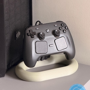 May include: A dark grey video game controller with dual analogue sticks, a directional pad, four action buttons labelled A, B, X, and Y, and two large touchpads, resting on a cream-coloured, oval-shaped, ribbed plastic stand with a matte finish.