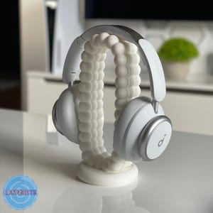May include: White headphone stand with a 3D printed design. The stand is shaped like a series of connected spheres. A pair of silver headphones are resting on the stand.