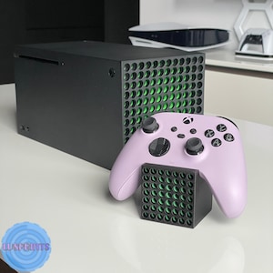May include: A black Xbox Series X console with a green grid pattern on the side. A purple Xbox controller is sitting on a black stand with a green grid pattern.