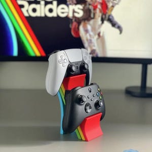 May include: A white and black PlayStation 5 controller and a black Xbox controller sit on a rainbow-coloured stand. The stand has red, yellow, green, and blue accents. The background shows the text "ARC Raiders" on a screen.