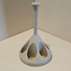 May include: A light grey golf tee holder, 3D-printed. It has a tall, slender stem and a flared base. The base has several oval openings for golf tees. The stem's top has a small, flat disc. The holder is approximately 15 cm tall.