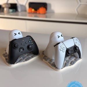 May include: Two white ghost-shaped controller stands with black eyes and mouths, each resting on a grey stone-textured base. One stand holds a black game controller, and the other holds a white game controller. A blue circular logo reads LUSPRINTS.