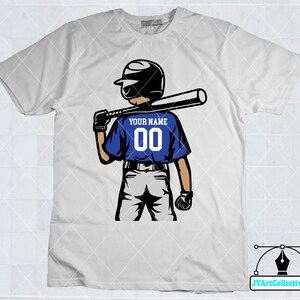 May include: White t-shirt with a blue baseball player graphic. The player is holding a bat and wearing a helmet. The text "YOUR NAME 00" is on the back of the player's jersey.