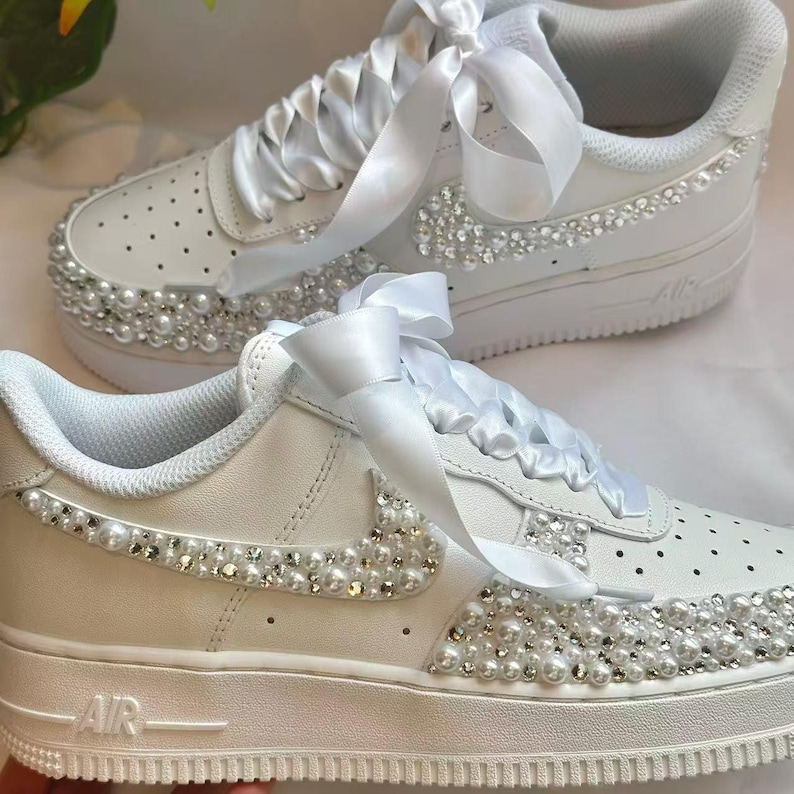 K&ouml;nnte beinhalten: Wei&szlig;e Turnschuhe mit Perlen- und Strassverzierungen. Die Schuhe haben wei&szlig;e Satinband-Schn&uuml;rsenkel und ein Perlen- und Strassdesign an den Seiten und Zehen. Das Wort "AIR" ist auf der Seite der Sohle sichtbar.