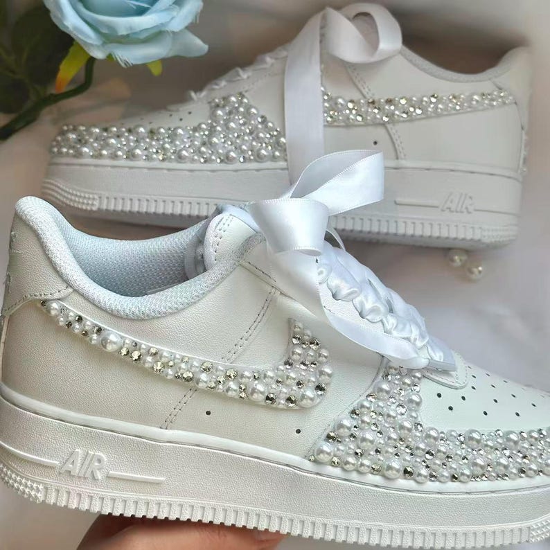 K&ouml;nnte beinhalten: Wei&szlig;e Turnschuhe mit Perlen und Strasssteinen verziert. Die Schuhe haben eine wei&szlig;e Sohle, ein wei&szlig;es Band zum Schn&uuml;ren und das Wort "AIR" auf der Seite. Eine blaue Blume ist im Hintergrund.