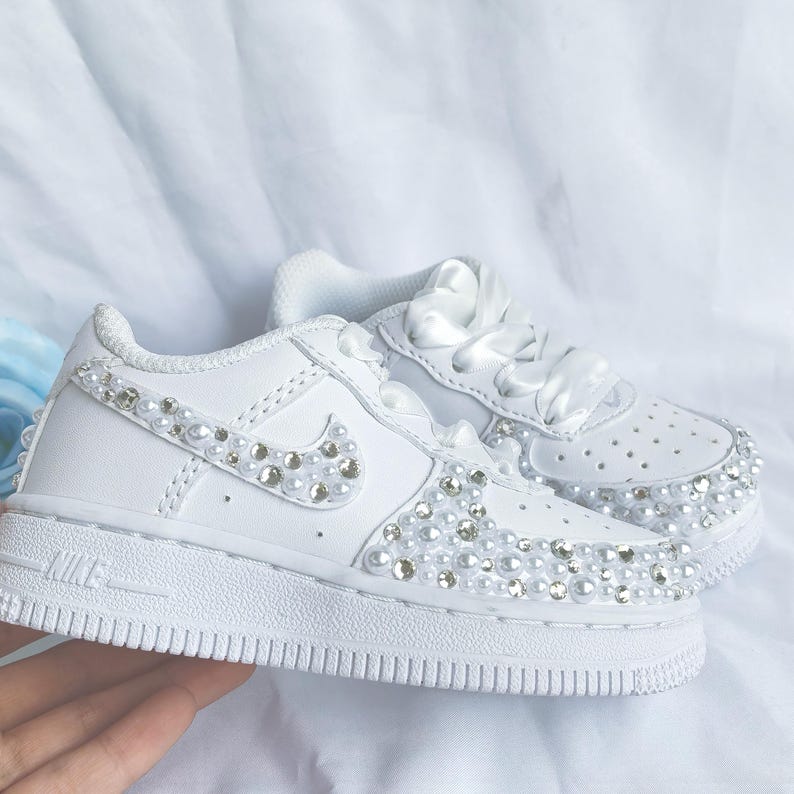 May include: White sneakers with pearl and rhinestone embellishments. The shoes feature a classic design with the Nike logo and satin ribbon laces. The toe and sides are adorned with pearls and rhinestones, adding a touch of glamour to the footwear.