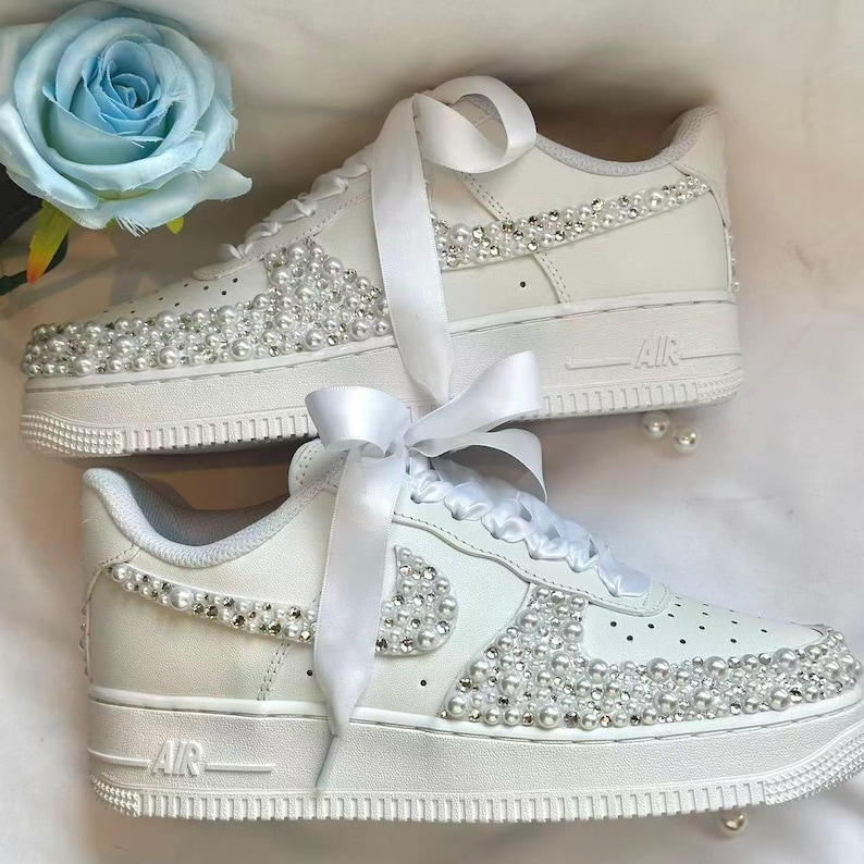 K&ouml;nnte beinhalten: Wei&szlig;e Turnschuhe mit Perlen und Strasssteinen verziert. Die Schuhe haben wei&szlig;e Satinband-Schn&uuml;rsenkel und Perlenakzente. Eine hellblaue Rose befindet sich im Hintergrund.