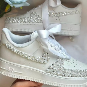 K&ouml;nnte beinhalten: Wei&szlig;e Turnschuhe mit Perlen und Strasssteinen verziert. Der Nike-Swoosh ist ebenfalls mit Perlen verziert. Die Schn&uuml;rsenkel wurden durch wei&szlig;e Satinb&auml;nder ersetzt. Die Schuhe haben ein klassisches, sportliches Design mit einem Hauch von Eleganz.