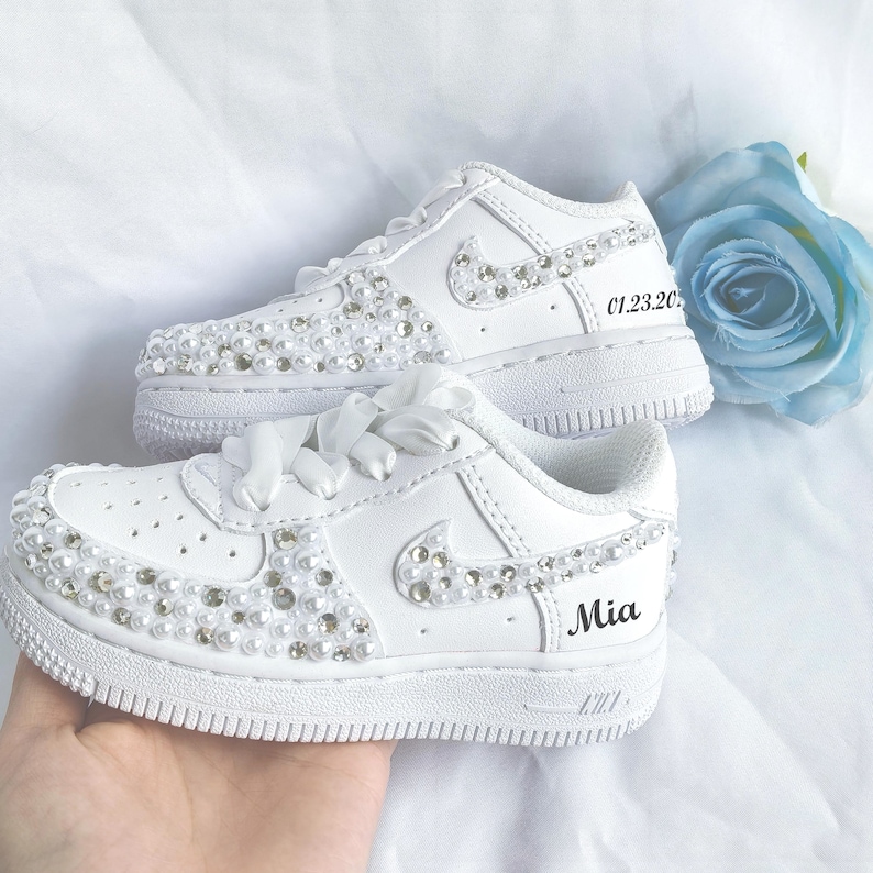 May include: White sneakers with pearl and rhinestone embellishments. The Nike swoosh is adorned with rhinestones. The name "Mia" and the date "01.23.20" are printed on the shoes. White satin ribbon laces complete the look.