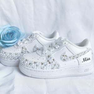 May include: White sneakers with pearl and rhinestone embellishments. The shoes have white satin laces and the name "Mia" is printed on the side. A light blue rose is in the background.
