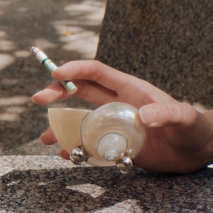 May include: A pearlescent seashell ashtray with silver accents. The ashtray is held in a hand, and a lit cigarette rests above it. The ashtray has a unique, natural shape and is designed to hold a cigarette.