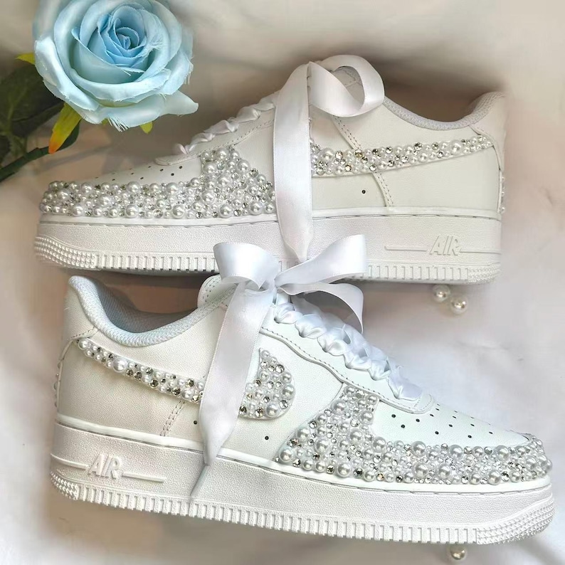 K&ouml;nnte beinhalten: Wei&szlig;e Turnschuhe mit Perlen und Strasssteinen verziert. Die Schuhe haben eine wei&szlig;e Sohle, ein wei&szlig;es Band und eine hellblaue Blume im Hintergrund. Diese Turnschuhe sind eine stilvolle und einzigartige Schuhoption.