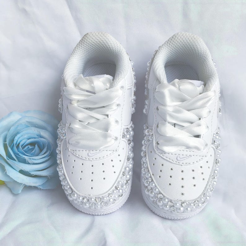 May include: A pair of white sneakers with pearl embellishments and satin ribbon laces. The shoes have a classic design with a perforated toe box and a small Nike logo. A light blue rose is visible in the background.