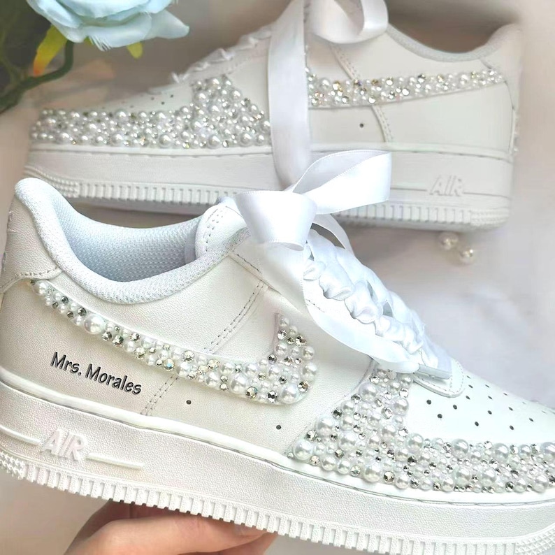K&ouml;nnte beinhalten: Wei&szlig;e Turnschuhe, verziert mit Perlen und Strasssteinen. Der Nike-Swoosh und der Zehenbereich sind mit glitzernden Akzenten verziert. Der Text "Mrs. Morales" ist auf der Seite aufgedruckt. Wei&szlig;e Satinb&auml;nder ersetzen die Schn&uuml;rsenkel, wodurch ein Brautthema entsteht.