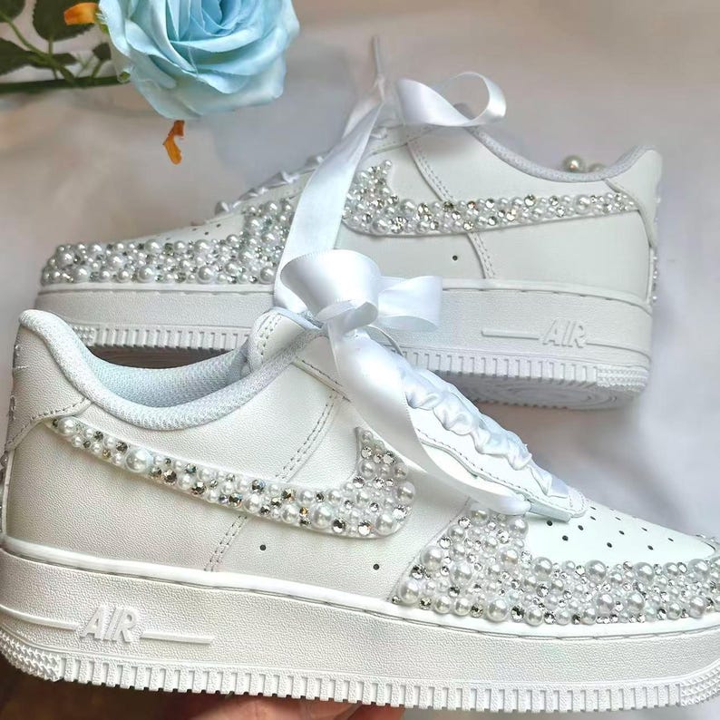 K&ouml;nnte beinhalten: Wei&szlig;e Turnschuhe mit Perlen und Kristallen verziert. Die Schuhe haben ein wei&szlig;es Band und sind mit glitzernden Akzenten verziert. Die Turnschuhe haben ein klassisches Design mit dem Wort "AIR" auf der Seite.