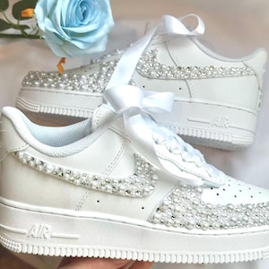 K&ouml;nnte beinhalten: Wei&szlig;e Sneaker mit Perlen und Strasssteinen verziert. Die Schuhe haben ein wei&szlig;es Band und den Nike-Swoosh. Die Zehen und Seiten sind mit dekorativen Elementen verziert, was einen glamour&ouml;sen Look erzeugt.