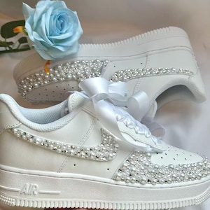 K&ouml;nnte beinhalten: Wei&szlig;e Turnschuhe, verziert mit Perlen und Strasssteinen. Die Schuhe haben ein wei&szlig;es Band und eine hellblaue Rose. Das Wort "AIR" ist auf der Seite sichtbar. Diese individuellen Turnschuhe sind ideal f&uuml;r besondere Anl&auml;sse.