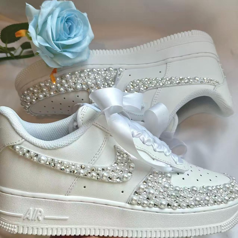 K&ouml;nnte beinhalten: Wei&szlig;e Turnschuhe, verziert mit Perlen und Strasssteinen. Die Schuhe haben ein wei&szlig;es Band und eine hellblaue Rose. Das Wort "AIR" ist auf der Seite sichtbar. Diese individuellen Turnschuhe sind ideal f&uuml;r besondere Anl&auml;sse.