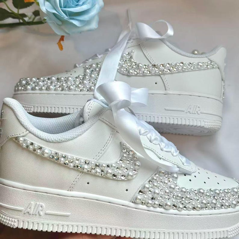 K&ouml;nnte beinhalten: Wei&szlig;e Turnschuhe mit Perlen und Strasssteinen verziert. Die Schuhe haben eine wei&szlig;e Schleife und sind mit einem Perlen- und Strassmuster auf dem Nike-Swoosh und im Zehenbereich verziert. Die Schuhe haben das Wort "AIR" auf der Seite.
