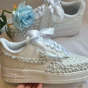 K&ouml;nnte beinhalten: Wei&szlig;e Turnschuhe mit Kunstperlen und Strasssteinen verziert. Die Schuhe haben eine Schleife aus Satinband und eine hellblaue Rose. Das Design umfasst den Nike-Swoosh und das Wort "AIR" auf der Sohle.