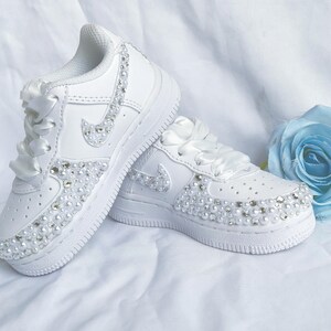 May include: White sneakers with pearl and rhinestone embellishments. The shoes feature a white ribbon lace-up design and a classic swoosh logo. The toe and side panels are adorned with decorative elements.
