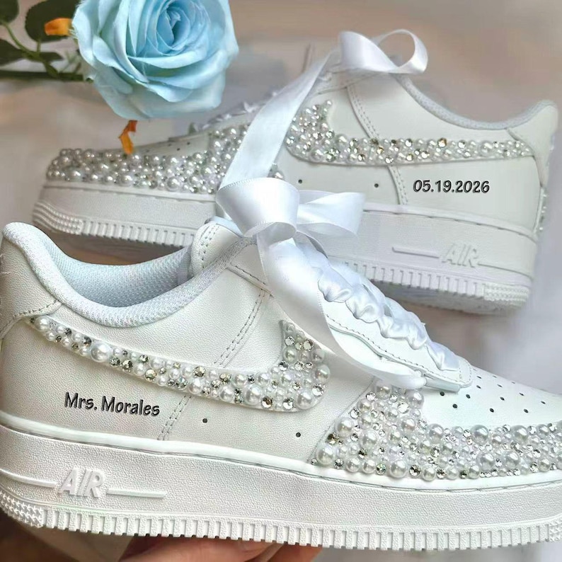 K&ouml;nnte beinhalten: Wei&szlig;e Turnschuhe mit Perlen und Strasssteinen verziert. Die Schuhe haben ein wei&szlig;es Band und die Texte "Mrs. Morales" und "05.19.2026". Die Turnschuhe haben ein klassisches Design mit wei&szlig;er Sohle und dem Wort "AIR" an der Seite.