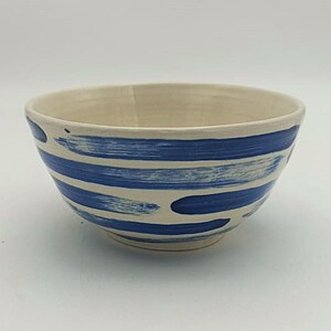 Puede incluir: Un tazón de cerámica blanco con pinceladas azules. El tazón tiene una forma redondeada y un acabado liso y brillante.