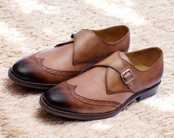 May include: A pair of brown leather monk strap shoes with a cap toe design. The shoes feature a buckle closure and decorative perforations along the edges. The leather has a gradient color, transitioning from light brown to a darker shade at the toe.