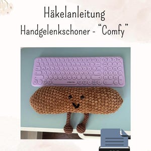 Puede incluir: Un reposamuñecas de ganchillo marrón con una cara sonriente y pequeñas patas está debajo de un teclado morado. El texto "Häkelanleitung Handgelenkschoner - Comfy" está en la parte superior. Un icono PDF está en la esquina inferior derecha.