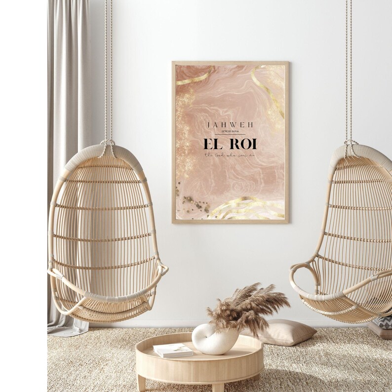 Op de afbeelding: Een ingelijste kunstdruk met de woorden "JAHWEH EL ROI" in zwarte tekst op een roze en gouden abstracte achtergrond. Twee hangende rieten stoelen en een ronde houten tafel met een vaas en decoratieve items staan op de voorgrond.