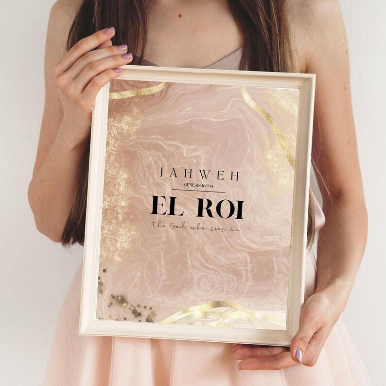 Op de afbeelding: Een ingelijste print met een zachtroze en gouden abstract ontwerp. De tekst op de print luidt "JAHWEH EL ROI" met de woorden "the God who sees me" eronder. De lijst is lichtbeige.